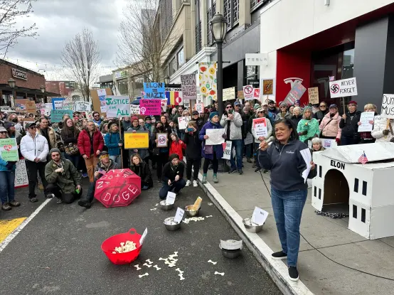 A crowd of hundreds stand in the road in front of a Seattle Tesla dealership. Congress woman Pramila Jayapal stands with the crowd holding a microphone