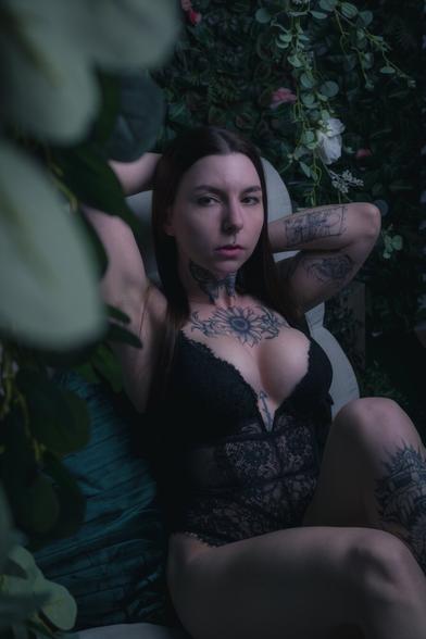 A woman with long dark hair and intricate tattoos lounges on a velvet chair surrounded by lush greenery and flowers. She wears a black lace bodysuit, her arms resting behind her head, gazing into the camera with a calm yet intense expression. The soft lighting and deep tones create a moody, ethereal atmosphere.