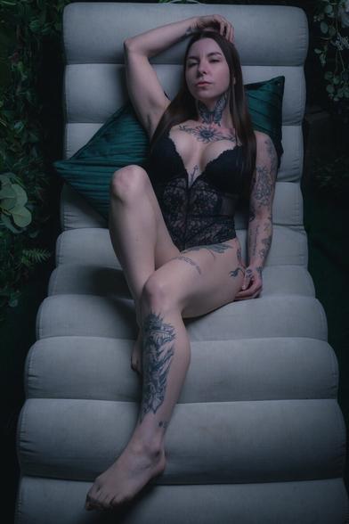 A woman with long dark hair and intricate tattoos lounges on a velvet chair surrounded by lush greenery and flowers. She wears a black lace bodysuit, her arms resting behind her head, gazing into the camera with a calm yet intense expression. The soft lighting and deep tones create a moody, ethereal atmosphere.