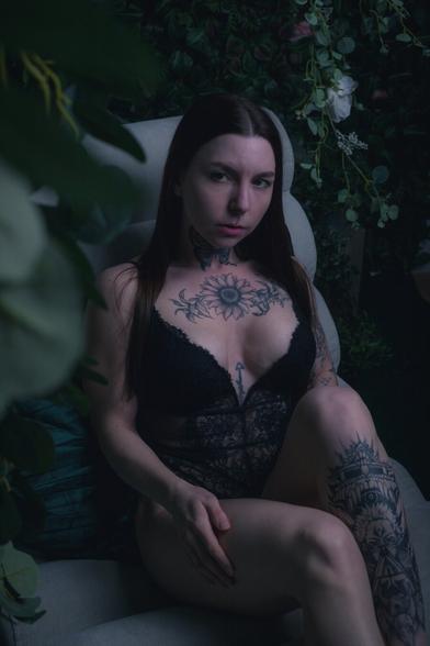 A woman with long dark hair and intricate tattoos lounges on a velvet chair surrounded by lush greenery and flowers. She wears a black lace bodysuit, her arms resting behind her head, gazing into the camera with a calm yet intense expression. The soft lighting and deep tones create a moody, ethereal atmosphere.