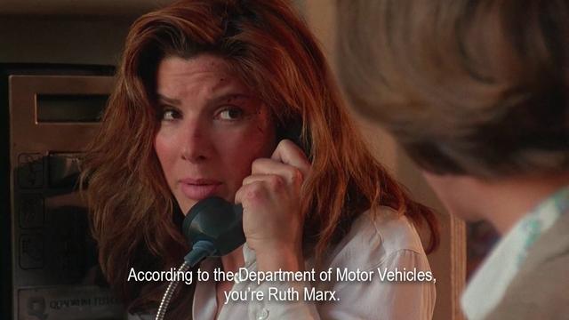 A woman with long hair is on a payphone in an American embassy. A white woman approaches her and asks her name. She says, "According to the Department of Motor Vehicles, you’re Ruth Marx.” The woman on the phone looks at her skeptically.