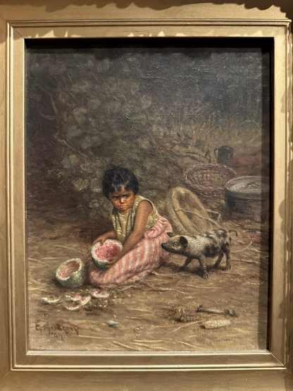 photo of painting in gold frame on display at museum: small oil portrait of a young Pomo child dressed in green and white striped top and pink and white striped bottom, sitting on ground holding sliced half of watermelon with other half and other rinds on ground, looking at a little spotted piglet who is eyeing the melon; Pomo baskets visible in background