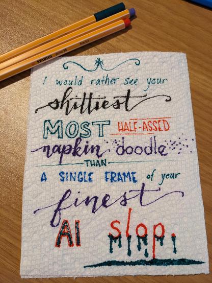 In various colored markers and in a mix of print, caps, and faux calligraphy, the text has been written on a half paper towel: "I would rather see your shittiest most half-assed napkin doodle than a single frame of your finest AI slop." There is a goo drawn dripping from the word "slop" and all the letters are a bit blurred and spread (as markers on paper towels are wont to do). The background is a wooden desk with some of the markers visible in the upper corners.