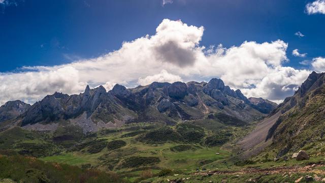 The image shows a scenic landscape with a range of mountains under a partly cloudy sky. The foreground features a grassy area with some rocky patches. The mountains are rugged and prominent, contributing to a dramatic and picturesque view.