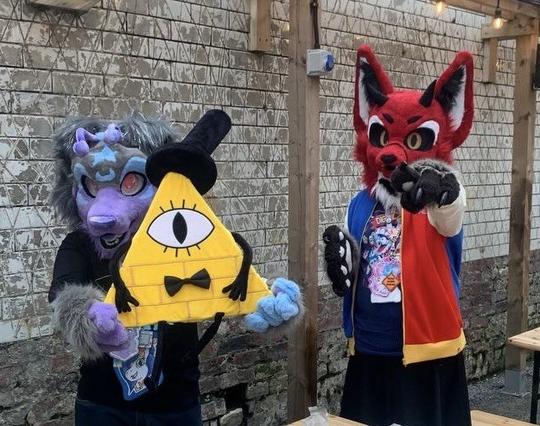 Midnight (alien dog fursuit) holds Bill Cipher while Lillith (devil fox fursuit) points at the camera.
