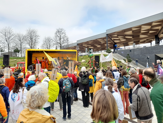 People gathered in front of a small stage with musicians in orange overalls. A person wearing a moth costume and a person in a grasshopper costume are dancing on stilts in front of the stage.