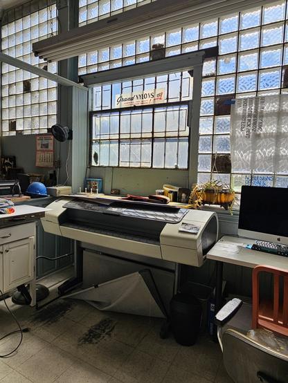 Photograph of the inside of  Noble Signs showing a wall of 1950's style glass bricks. Infront of the glass a huge printer, adesk with an apple branded monitor. To the lect we see a blue co struction sire helmet and old style walk calendar