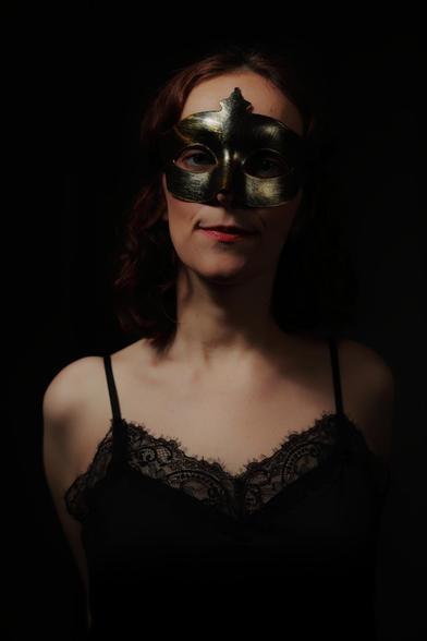 The image shows a person wearing a decorative mask. The mask is metallic and covers the upper part of the face. The person is dressed in a dark, lace-trimmed garment with thin straps. The background is dark, highlighting the subject.