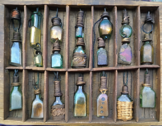 A charmingly irregular wooden typecase style compartmented box, holding an eclectic collection of rustic glass bottles with a handmade look, with a variety of magical contents.