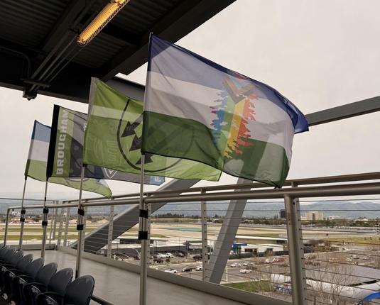 Four flags flying at the top of the visitor’s supporters section. Cascadia flag in LGBTQ+ colors. Iron Front. Emerald City Supporters. Cascadia flag with a slice of pizza.