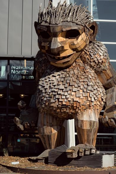 a sculpture of a troll made of wooden scraps faces the camera. The figure is about 20 feet tall. It is standing in front of the Nordic Museum