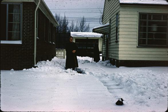 On the left, a red brick house. On the right, a yellow wood paneled house. In the middle a shared drive way mostly covered in about six inches of snow. Two older women in conservative black coats pose with their snow shovels.