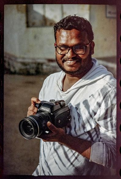 A color photo of a man smiling at the camera and holding a medium format camera.