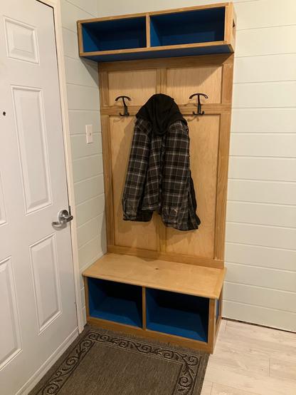 A cost rack with three hooks. Maple and oak with cubbies for hats and shoes in blue. A plaid hoody hangs on the centre hook