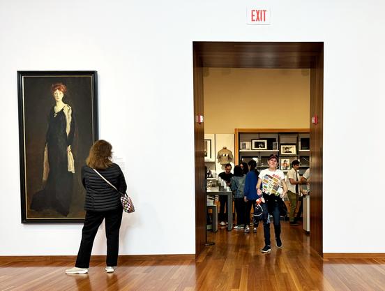 On the right side a person in a white shirt walks through a doorway from a crowded gift shop. On the left is a painting of a white woman in a black dress. A person in a black shirt, black pants, white shoes and a white purse is standing to the right of the painting looking toward the doorway.