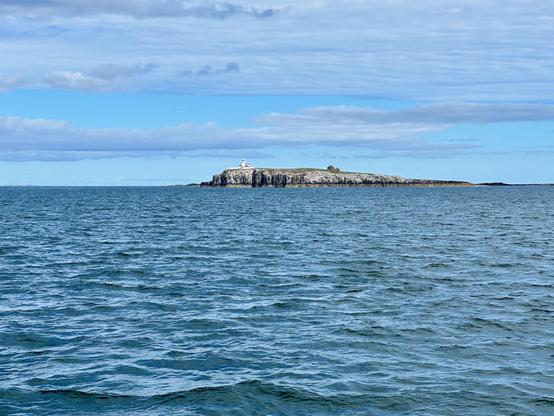 The image shows a distant, rocky island with a lighthouse on it, surrounded by a body of water. The sky is partly cloudy with shades of blue and white. The water appears calm, with gentle waves and reflections of the sky. The island is mostly barren, with some vegetation visible near the lighthouse.