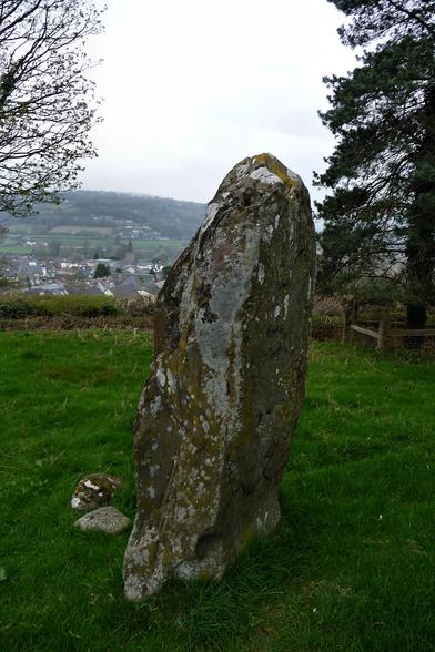 An upright slab of stone, over 6 feet tall, standing in a grass field with trees on either side. The setting is elevated over a valley, and the houses and church spire of small town can be seen in the valley below. Wooded hills rise beyond that. It's a bit grey and overcast.