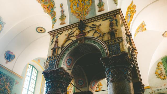 The central podium in the Łańcut Synagogue in Łańcut, Poland. A square podium with decorative arches opening on all four sides. Intricate textures, shapes, Hebrew writing and designs decorate the arch and surrounding synagogue. Colors of gold, greens, deep blue, yellow, lime, pink. A window in the background lets in bright sunlight, illuminating the room.