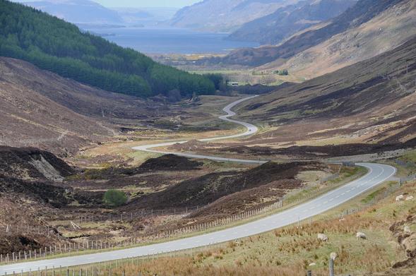 Glen Docherty. The image shows a view down the glen with the road sweeping broadly from side to side. There’s part of a forest on the left in the middle distance and a large loch flanked by mountains at the top of the frame.