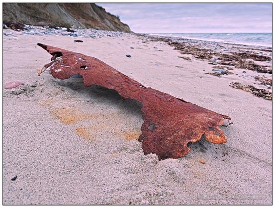 Bodennahe Weitwinkelaufnahme eines komplett rötlich verrosteten, halbrund gebogenen und an den Rändern bereits ausgefressenen Blechs im Sand des Ostseestrandes, im Hintergrund links das Steilufer, rechts das Meer.