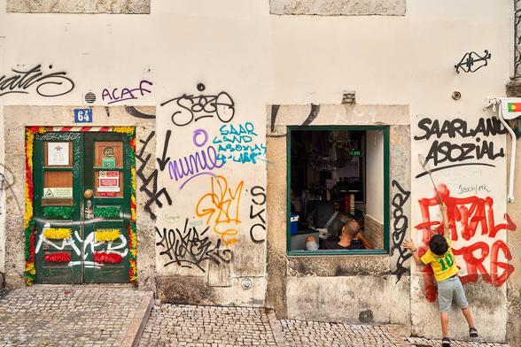 Street scene. Café in Lisbon. Yellow-white wall scribbled full with meaningless (and ugly) graffitti. To the left is a green door that is closed adorned with ribbons in the Portuguese colours. To the right is a window where is a kitchen with a man sitting facing the interior. Still right is a boy with green-grey short-pants and a bright yellow shirt with a badminton bat chasing flies.