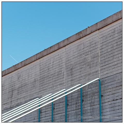Kahle Betonmauer vor blauem Himmel mit fünf daran befestigten metallenen Streben einer Tragekonstruktion; der Aufbau dieser streng grafischen Bildkomposition wird von links unten nach rechts oben verlaufenden Diagonalen bestimmt.