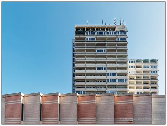 70er-Jahre Betonhochhaus vor blauem Himmel in der rechten Bildhälfte, waagerecht im unteren Bildviertel quer durchs Bild verlaufender Ausschnitt einer verwinkelten Betonmauer, deren senkrechte Abschnitte abwechselnd in Betongrau und einem zarten, pastelligen Rosa gehalten sind. Modulierenden Lichtreflexe, weich gestreut vom verwinkelten Mauerwerk, geben der Mauer optisch Tiefe.