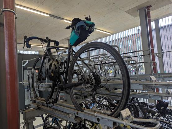 Fahrrad in auf der oberen Etage eines 2 stöckigen Fahrradständer.

Im Hintergrund sieht man noch weitere Fahrräder die unten stehen und das Gitter als Einbruchschutz.