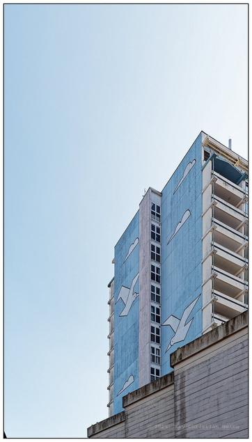 70er-Jahre Betonhochhaus in schmalem Hochformat vor blauem Himmel fotografiert. Die Fassade ist in einem zarten, pastelligen Hellblau ausgeführt, auf das in einfachem Stil weiße fliegende Möwen und Wolken gemalt sind. Im rechten unteren Bildviertel ist der Ausschnitt einer verwinkelten Betonmauer zu sehen