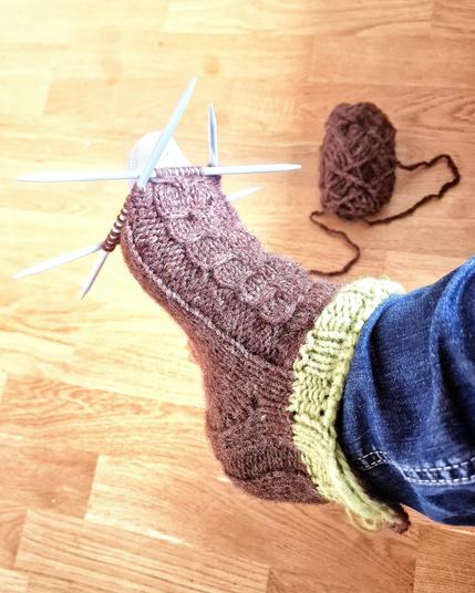 En brunt socka med pistagegrönt kort skaft som är nästan färdigstickad som prövas på en högerfot med fyra strumpstickor ännu i I framkant på stickningen. 

Benet har blå jeans och golvet är ett parkettgolv.