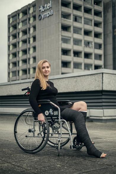 Junge Frau sitzt mit zwei Gipsbeinen in einem Rollstuhl in brutalistisch urbaner Umgebung