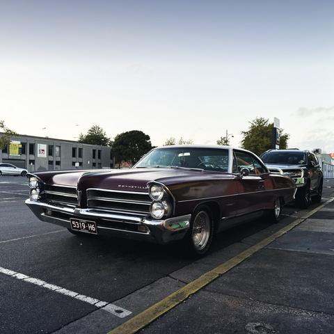 The front view from the left-hand front corner of an old two-door and very long car, parked next to a pavement - the car is called a “Bonneville”. Two headlights in a vertical configuration on each side, and a slightly pointy nose in the middle. It’s a lovely dark red, and a clear early morning sky is behind, with a couple of trees and a distant building.
