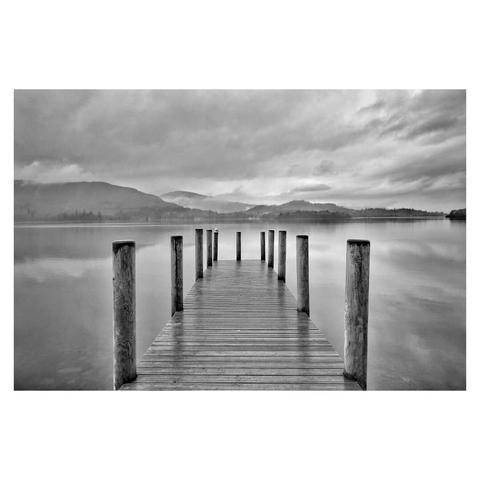 This is a black and white landscape format image of a jetty pointing out across a lake. The viewer looks straight down the jetty into the centre of the image. In the distance mountains are shrouded in clouds. The water of the lake reflects the mountains and the clouds. Taken in the Lake District with a Sony RX100.