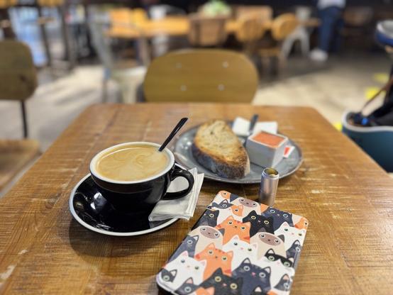 Mesa de madera recia con el Kobo, la tacita negra del café súper rico con leche fría de avena y el plato con la tostada de aceite y tomate. Por encima está el tubito donde tengo la pastilla de la estúpida diabetes.