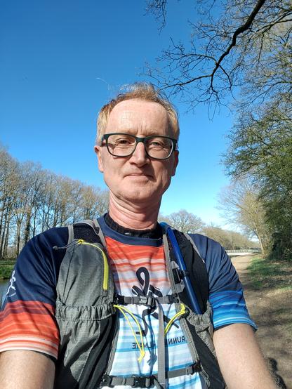 Selfie of runner in the sun alongside a canal. Trees in the side, blue sky above. Runner is wearing climate striped running shirt and a camelbag.