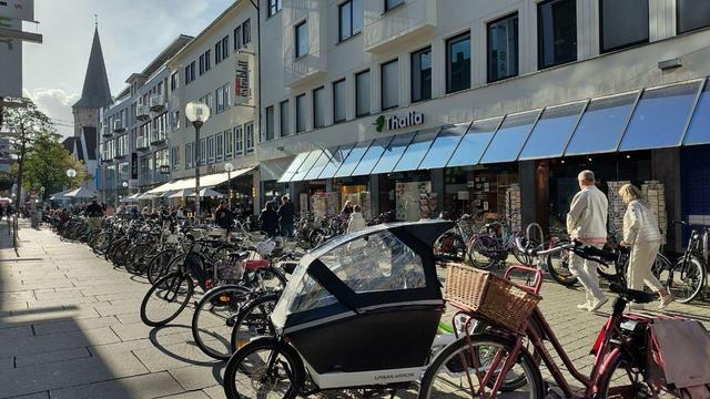Das Bild zeigt eine belebte Fußgängerzone in Osnabrück. Es gibt viele Fahrräder, die am Rand geparkt sind. Entlang der Zone stehen Gebäude mit Geschäften und Wohnungen. Im Hintergrund ist ein Kirchturm zu sehen. Das Bild ist bei Tageslicht aufgenommen und wirkt sonnig.