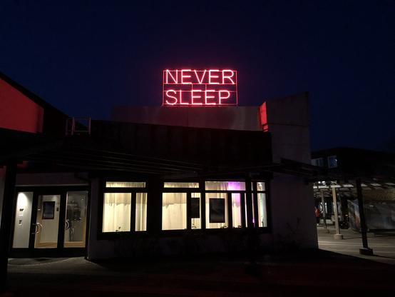 "Never Sleep" written in neon letters, on the roof of a low part of the Kulturhaus of Lüdenscheid. Night time, sky still glows a faint blue hue.