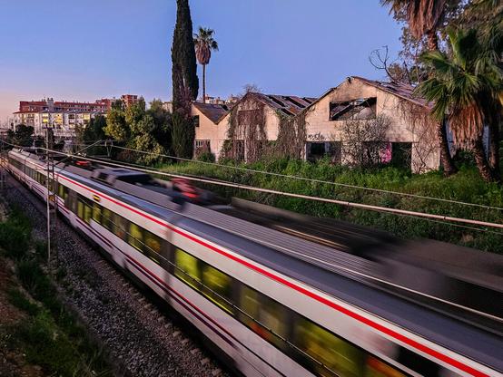 A white train with red stripes passing through a cutting, blurred due to its speed. In the background are derelict warehouses and the early morning sky.
