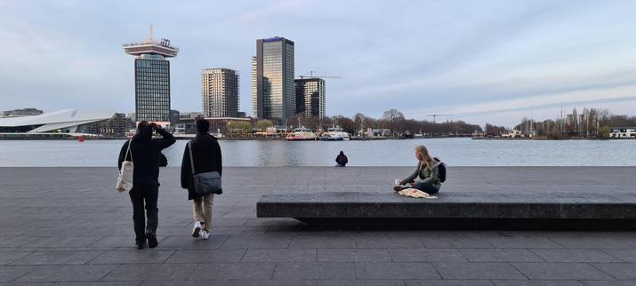 A landscape oriented photo of the grey stone paved quay at the river Amstel behind Amsterdam Central Station. In the foreground a couple walking past a young woman sitting on a long,  stone slab bench holding a coffee. Visible between them there's someone sitting at the water's edge. In the background the Amsterdam north skyline with the Eye cinema, Amsterdam Tower and three highrises among the trees lining the shore against a blue but cloudy sky.