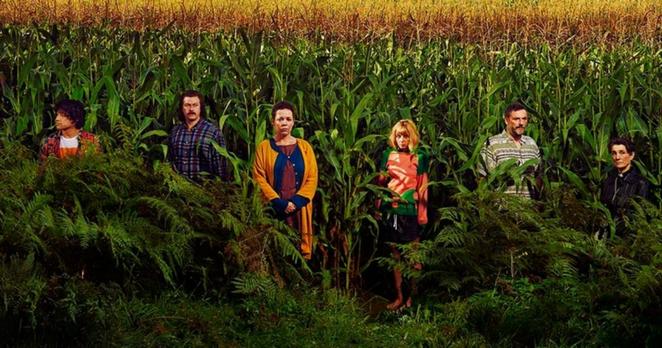 still image from the english series, 'flowers', shows the eponymous family (including shun, the illustrator) stood on the edge of mrs beckett's cornfield. 
from L to R: will sharpe, daniel rigby, olivia colman, sophia di martino, julian barratt & harriet walker...