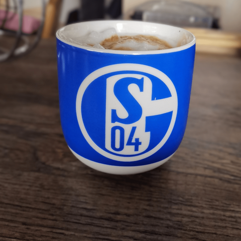 Eine Bauchige königsblaue Tasse mit dem Logo des FC Schalke 04.
Die Tasse steht auf einem dunklen Holzuntergrund