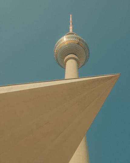 The image shows the Berlin TV Tower (Fernsehturm) viewed from a dramatic low angle. The tower's distinctive spherical observation deck with its metallic, reflective surface is prominently visible at the top of a tall white concrete shaft. In the foreground, there's a geometric, triangular architectural element, likely part of a nearby building or structure, creating an interesting compositional contrast with the tower. The background features a clear blue sky, highlighting the tower's iconic silhouette against it. This landmark is one of Berlin's most recognizable structures and a symbol of the city.
