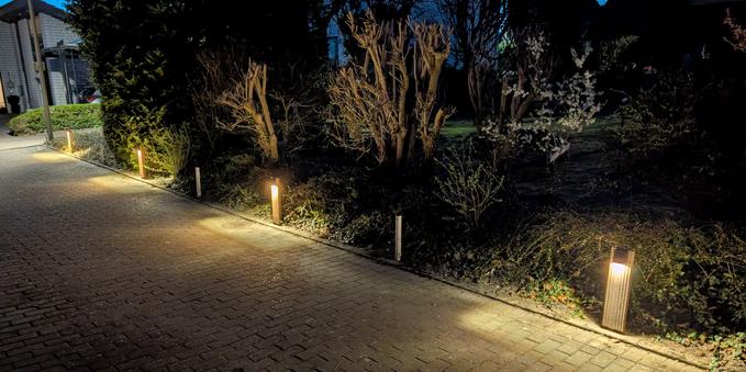 Entlang eines gepflasterten Hofes, welcher nach rechts mit einer Hecke abschließt stehen vor der Hecke vier kleinen Bodenstrahler und erzeugen im Dämmerlicht rechteckige Lichtflächen auf dem Pflaster.
