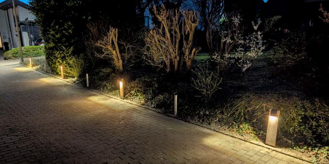 Entlang eines gepflasterten Hofes, welcher nach rechts mit einer Hecke abschließt stehen vor der Hecke vier kleinen Bodenstrahler und erzeugen im Dämmerlicht rechteckige Lichtflächen auf dem Pflaster.