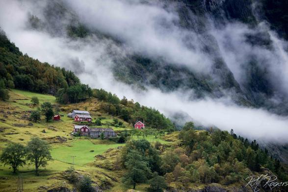 Photograph of a mountain farm perched on a green cliff in Norway's Aurlandsfjord. A morning mist is slowly burning off.