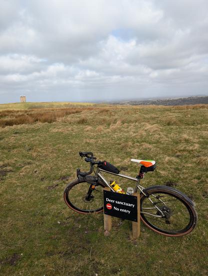 This image shows a bicycle leaning against a "Deer sanctuary No entry" sign in a wide, open landscape. The bike is a gravel bike with a titanium frame. The sign is in the foreground, and the landscape stretches out to a distant horizon with a cloudy sky. The ground is a mix of green grass and brown patches, suggesting a moorland or hilltop setting. In the distance, a small building  is visible.
