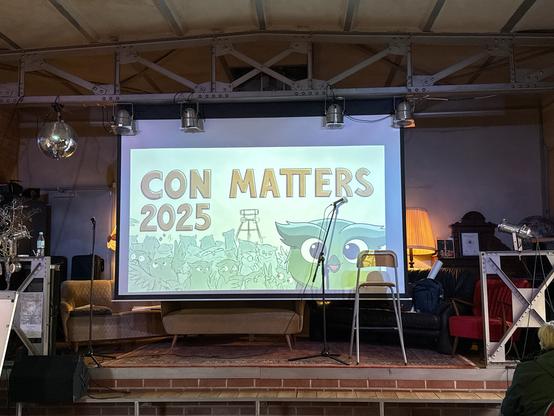 Foto einer leeren Bühne,  auf der eine große Leinwand zu sehen ist, auf die „Con Matters 2025“ mit einer Ansammlung einer Reihe von cartoonhaften Eupen zu sehen ist - ebenso ein Förderturm im Hintergrund  eine größere Eule im Vordergrund schaut in unsere Rochtung. 
Außerdem Stühle, Mikrofons.