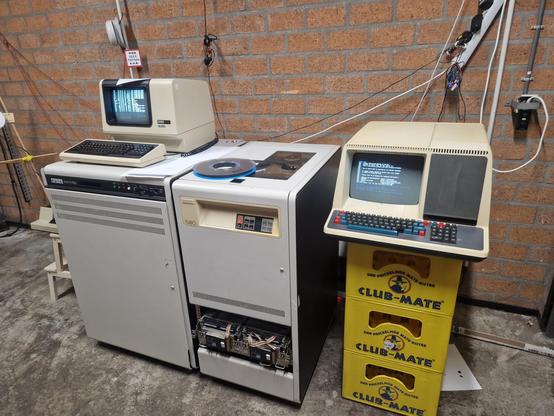 Row of historical computer equipment: Hp7475A plotter, VAX-11/750 with a VT100 terminal atop it, TU80 tape drive with two fujitsu 8" harddrives below it, VT55 terminal resting on a stack of club mate crates.