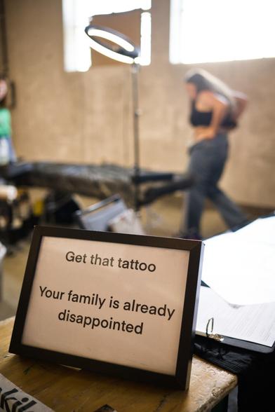A sign in the front says "get that tattoo, your family is already disappointed" while in the background you see a girl out of focus getting ready to get inked