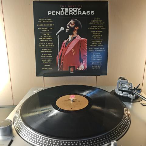 A vinyl record sits on a turntable. Behind the turntable, a vinyl album outer sleeve is displayed. The front cover shows Teddy Pendergrass singing in to a microphone.